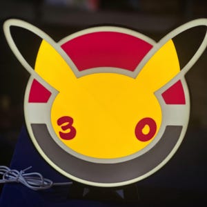 May include: A yellow, red, and black circular light-up sign featuring a cartoon character's face. The number "3" and a red circle are visible. The sign has a white border and a black base.