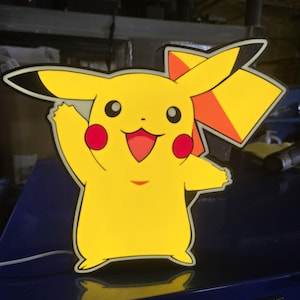 May include: A yellow Pikachu-shaped light with a black outline. The light features Pikachu's iconic red cheeks, a smiling mouth, and a waving arm. The light has a black and orange accent on the ear. A white cord extends from the bottom.