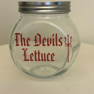 May include: A clear glass jar with a silver lid. The jar has the words "The Devils Lettuce" in red gothic lettering with a red trident symbol. The jar is empty and sits on a white surface.