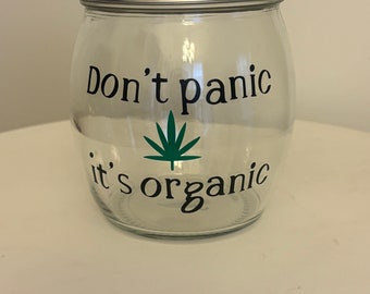 May include: A clear glass jar with a silver lid. The jar has a green marijuana leaf and the text "Don't panic it's organic" printed on it.
