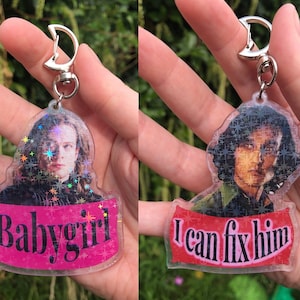 May include: Two clear plastic keychains with a holographic finish. The keychains feature a portrait of a man with long hair. The text "Babygirl" is on one keychain and "I can fix him" is on the other keychain.