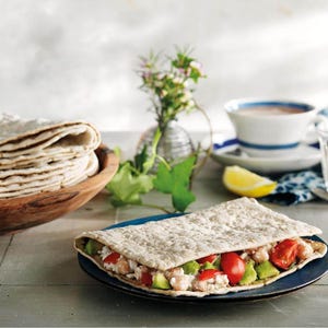 May include: A flatbread sandwich filled with avocado, tomatoes, and a white filling, served on a blue plate. A stack of flatbreads sits in a wooden bowl. A cup of soup and a lemon wedge are in the background.