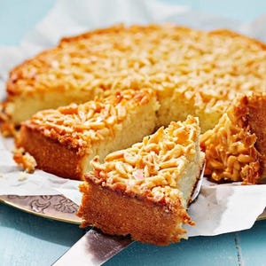 Puede incluir: Un pastel de almendras parcialmente cortado sobre un plato decorativo. El pastel está cubierto de láminas de almendras tostadas. Una porción se levanta con un servidor plateado, revelando el interior húmedo del pastel.
