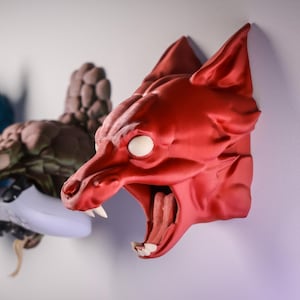 Może przedstawiać: Trzy dekoracyjne rzeźby ścienne przedstawiające mityczne stworzenia. Pierwsza to niebieska głowa smoka, druga to brązowe stworzenie, a trzecia to czerwona głowa wilka. Każda ma białe oczy i jest zamontowana na białej ścianie.