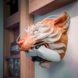 K&ouml;nnte beinhalten: Eine Wandskulptur eines Tigerkopfes in Orange und Wei&szlig;, die einen wei&szlig;en und schwarzen Gamecontroller h&auml;lt. Die Skulptur ist an einer hellen Wand befestigt, mit Holzregalen im Hintergrund.