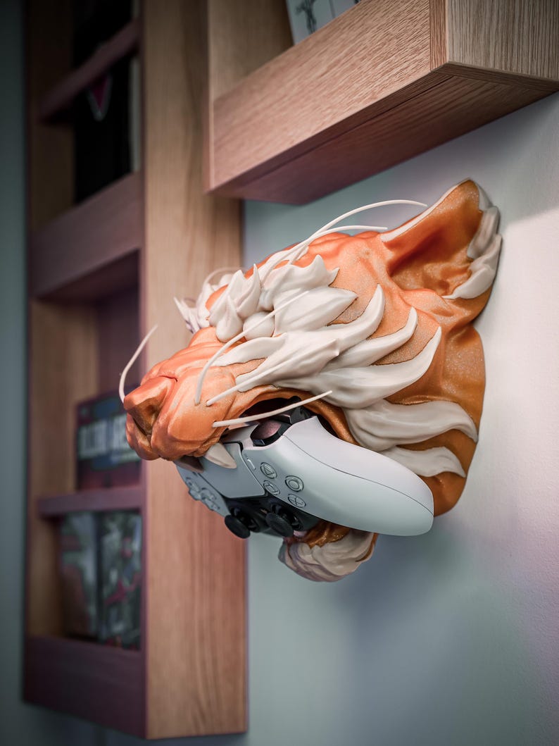 K&ouml;nnte beinhalten: Eine Wandskulptur eines Tigerkopfes in Orange und Wei&szlig;, die einen wei&szlig;en und schwarzen Gamecontroller h&auml;lt. Die Skulptur ist an einer hellen Wand befestigt, mit Holzregalen im Hintergrund.