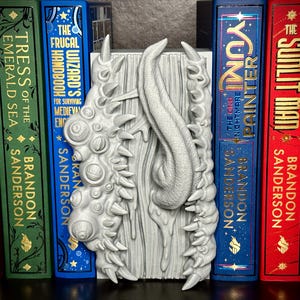 Puede incluir: Un sujetalibros gris de fantas&iacute;a con un dise&ntilde;o texturizado, con un elemento central en forma de lengua y protuberancias puntiagudas. Se encuentra entre varios libros de tapa dura, incluyendo "The Sunlit Man" y "Tress of the Emerald Sea".