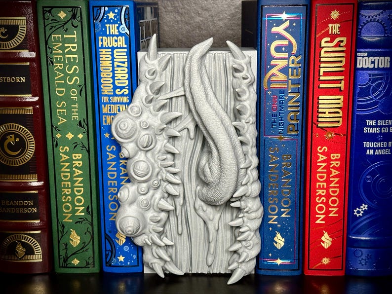 Puede incluir: Un sujetalibros gris de fantas&iacute;a con un dise&ntilde;o texturizado, con un elemento central en forma de lengua y protuberancias puntiagudas. Se encuentra entre varios libros de tapa dura, incluyendo "The Sunlit Man" y "Tress of the Emerald Sea".