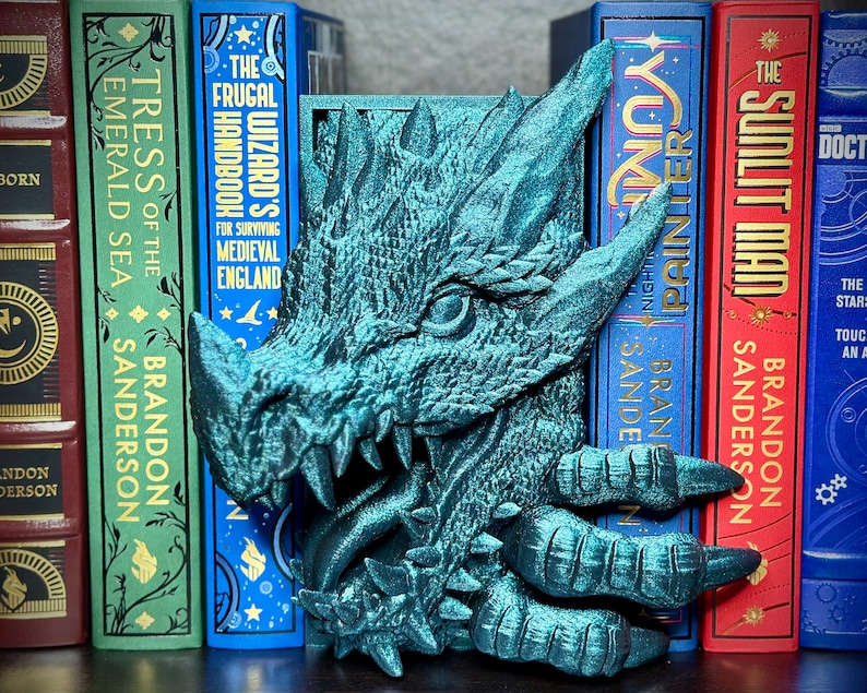 Puede incluir: Dos sujetalibros de drag&oacute;n verde turquesa con un acabado texturizado. La cabeza del drag&oacute;n est&aacute; mirando hacia adelante con la boca abierta y las garras extendidas. Los sujetalibros est&aacute;n colocados en una estanter&iacute;a con varios libros detr&aacute;s de ellos. Los libros tienen t&iacute;tulos que incluyen "The Frugal Wizard's Handbook", "Tress of the Emerald Sea", "Sunlit Man", y "Yume No Painller".