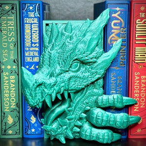 Puede incluir: Un sujetalibros de drag&oacute;n verde con un dise&ntilde;o detallado y texturado. La boca del drag&oacute;n est&aacute; abierta, mostrando dientes afilados. El sujetalibros est&aacute; sosteniendo dos libros, uno azul y otro rojo.