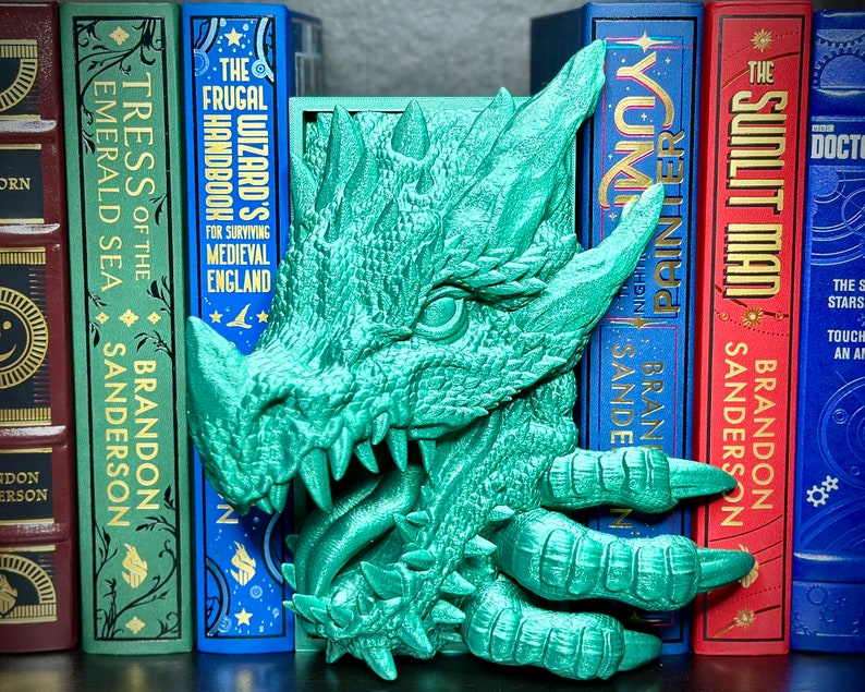 Puede incluir: Un sujetalibros de drag&oacute;n verde con un dise&ntilde;o detallado y texturado. La boca del drag&oacute;n est&aacute; abierta, mostrando dientes afilados. El sujetalibros est&aacute; sosteniendo dos libros, uno azul y otro rojo.