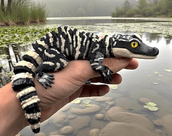 May include: A black and white patterned reptile with yellow eyes is held in a hand. The reptile has a detailed design, with stripes and spots. The background shows a lake with lily pads and a misty, overcast sky.