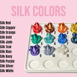 K&ouml;nnte beinhalten: Eine Pr&auml;sentation von elf kristallf&ouml;rmigen Objekten in verschiedenen Farben, darunter Rot, Kupfer, Orange, Gold, Jade, T&uuml;rkis, Blau, Marine, Lila, Silber und Wei&szlig;. Der Text "SILK COLORS" steht oben, mit nummerierten Farbbezeichnungen darunter.