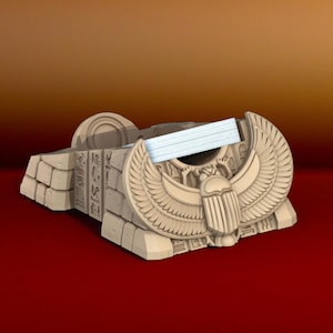 Puede incluir: Un dispensador de cinta de cerámica beige con forma de tumba egipcia con un diseño de escarabajo. El dispensador tiene un rollo de cinta blanco en su interior.