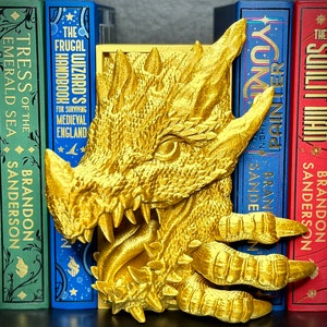 Puede incluir: Un sujetalibros de drag&oacute;n dorado con un dise&ntilde;o detallado. El drag&oacute;n est&aacute; sosteniendo un libro en sus garras. El sujetalibros est&aacute; sentado en un estante con una fila de libros detr&aacute;s.