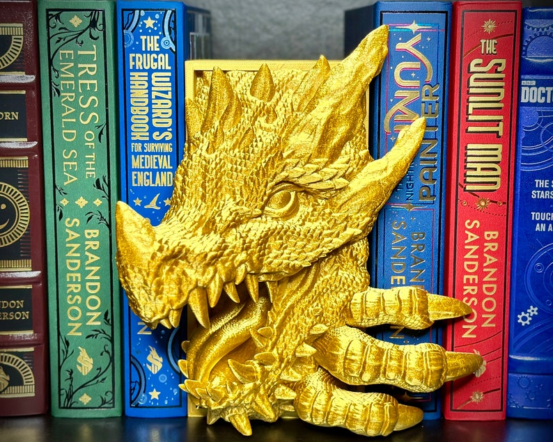Puede incluir: Un sujetalibros de drag&oacute;n dorado con un dise&ntilde;o detallado. El drag&oacute;n est&aacute; sosteniendo un libro en sus garras. El sujetalibros est&aacute; sentado en un estante con una fila de libros detr&aacute;s.