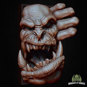 May include: A detailed 3D printed model of a snarling orc face with a large mouth and sharp teeth. The model is painted in a brown and gray color scheme and is mounted on a wooden base.