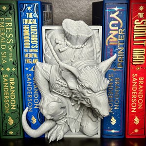 May include: A detailed, 3D-printed bookend featuring a stylized wolf-like creature with a bag and armor. The bookend is white and stands between several hardcover books with titles like "Tress of the Emerald Sea" and "The Frugal Wizard's Handbook".