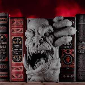 May include: A gray, 3D printed bookend shaped like a monster's head with a large mouth and sharp teeth. The bookend is placed between a row of six books with gothic and horror themed covers.