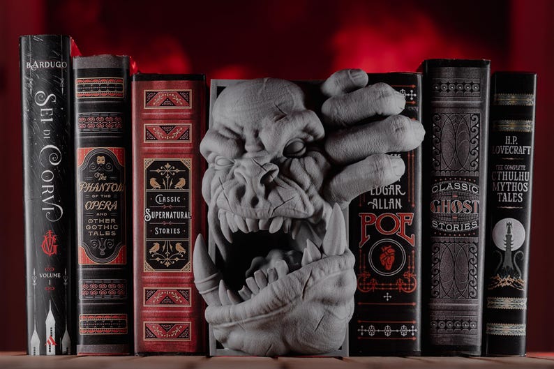 May include: A gray, 3D printed bookend shaped like a monster's head with a large mouth and sharp teeth. The bookend is placed between a row of six books with gothic and horror themed covers.
