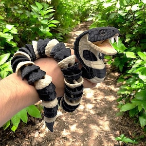 May include: An articulated snake toy in black and white, with an open mouth displaying white fangs. The snake's body is made up of alternating black and white segments. The toy is held against a blurred green backdrop of leaves.