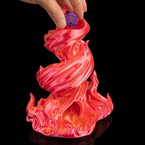 Puede incluir: Un soporte para dados en forma de llama roja y naranja con un dado de veinte caras morado y negro que se está colocando dentro. El soporte tiene un acabado brillante y está sobre un fondo negro.