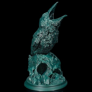 May include: A teal-colored sculpture of a raven perched atop a skull. The raven has an open beak, and the skull has a hollowed-out eye socket. The sculpture is on a round base and set against a black background.