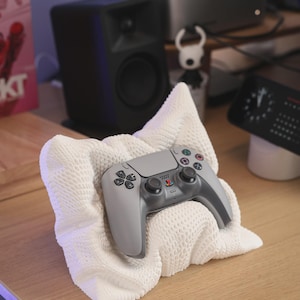 May include: A gray and black video game controller rests on a white, textured cloth. The controller has multiple colored buttons and directional controls. The cloth is folded and placed on a wooden surface.