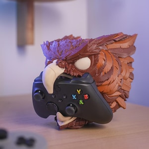 May include: A brown and tan owl figurine holding a black video game controller. The owl has detailed feather textures and white eyes. The controller has coloured buttons and directional controls. The figurine is on a wooden surface.