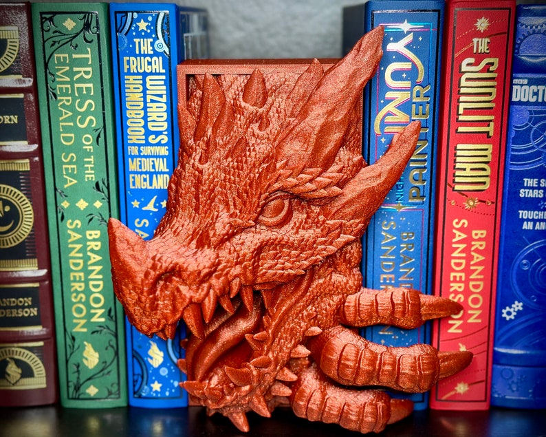 Puede incluir: Un sujetalibros de drag&oacute;n impreso en 3D en color cobre. El drag&oacute;n est&aacute; mirando hacia la izquierda y tiene la boca abierta. El sujetalibros est&aacute; sentado en un estante con libros detr&aacute;s.