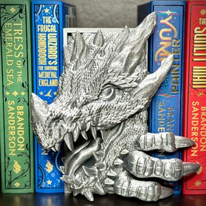 Puede incluir: Un sujetalibros de drag&oacute;n plateado con un dise&ntilde;o detallado y texturado. El drag&oacute;n est&aacute; sosteniendo un libro en sus garras y tiene la boca abierta, revelando dientes afilados. El sujetalibros est&aacute; sentado en un estante con una fila de libros detr&aacute;s.