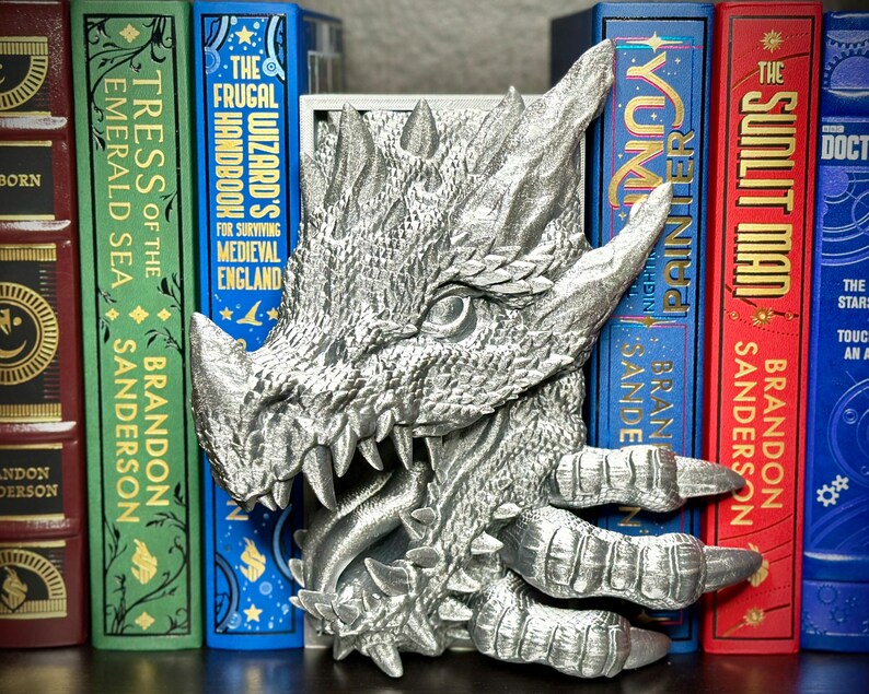 Puede incluir: Un sujetalibros de drag&oacute;n plateado con un dise&ntilde;o detallado y texturado. El drag&oacute;n est&aacute; sosteniendo un libro en sus garras y tiene la boca abierta, revelando dientes afilados. El sujetalibros est&aacute; sentado en un estante con una fila de libros detr&aacute;s.