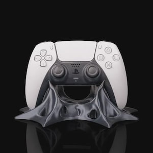 May include: A white and black PlayStation 5 controller resting on a dark gray, stylized stand. The controller features a white faceplate with black accents and the PlayStation logo. The stand has a unique, geometric design.