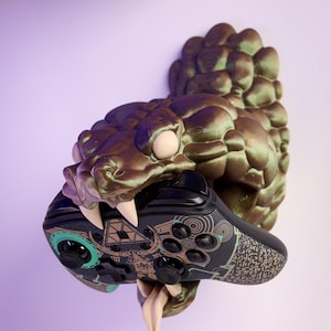 May include: A bronze-colored snake head wall mount holding a black and teal video game controller. The controller has a patterned design. The snake's fangs and tongue are visible.