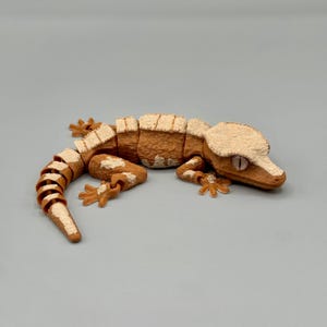 May include: A detailed, segmented gecko figurine in shades of brown and cream. The reptile's body is a warm brown, with cream-colored accents on its head, feet, and the edges of its body segments. The gecko is posed on a neutral gray surface.