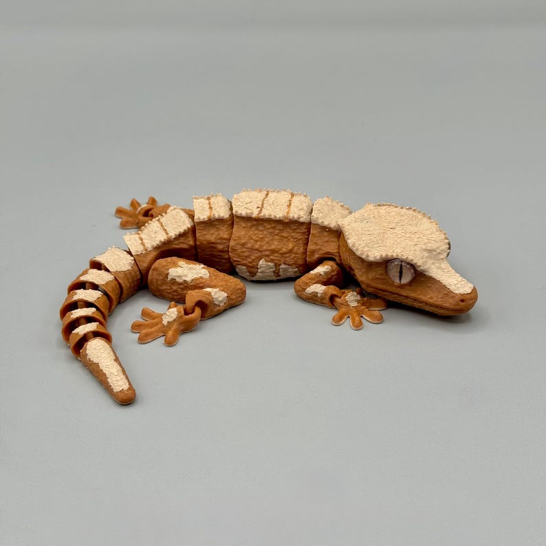 May include: A detailed, segmented gecko figurine in shades of brown and cream. The reptile's body is a warm brown, with cream-colored accents on its head, feet, and the edges of its body segments. The gecko is posed on a neutral gray surface.