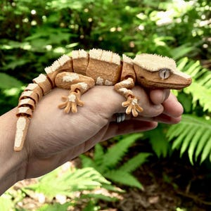 Pode incluir: Figura de gecko articulada e detalhada em tons de castanho e creme, com uma aparência texturizada. O gecko está posicionado em uma mão, mostrando seu design intrincado e características realistas. O fundo é um verde exuberante, criando um ambiente natural.