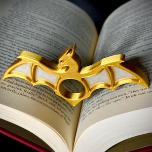 Puede incluir: Un anillo de dragón de color dorado con alas blancas. El anillo está apoyado en un libro abierto.