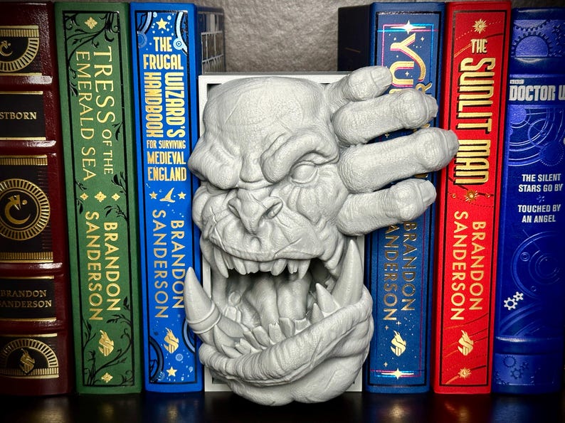 May include: A gray, 3D printed bookend featuring a snarling monster with a large, open mouth and a hand reaching out from the side. The bookend is placed between a set of books with gold lettering on the spines.