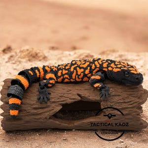 May include: A Gila monster figurine in orange and black, lying on a piece of driftwood. The lizard's body is segmented, with orange blotches and stripes on a black base. The figurine has small, detailed black feet and a black head.
