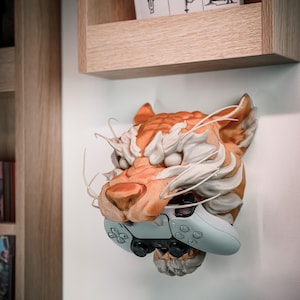 K&ouml;nnte beinhalten: Wandmontierte Tigerkopfskulptur in Orange und Wei&szlig;, die einen hellblauen Gamecontroller h&auml;lt. Die detaillierte Skulptur ist an einer wei&szlig;en Wand befestigt. Dar&uuml;ber befindet sich ein Holzregal.