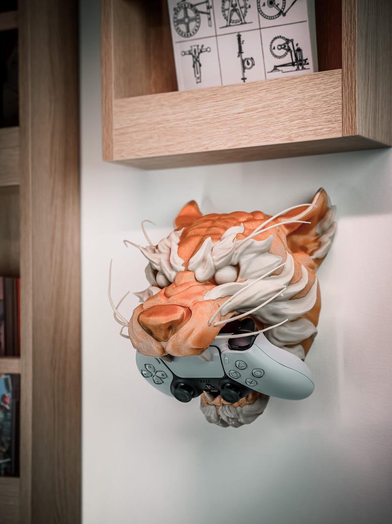 K&ouml;nnte beinhalten: Wandmontierte Tigerkopfskulptur in Orange und Wei&szlig;, die einen hellblauen Gamecontroller h&auml;lt. Die detaillierte Skulptur ist an einer wei&szlig;en Wand befestigt. Dar&uuml;ber befindet sich ein Holzregal.