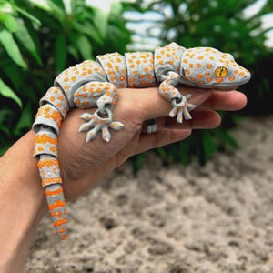 May include: A grey and orange articulated gecko toy with orange spots. The gecko is held in a hand, showing its flexible design. The toy's segmented body and limbs allow for various poses, making it a fun and interactive item.