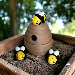May include: A brown, beehive-shaped decorative item with a round opening, topped with a yellow, black, and white bee figurine. Two additional bee figurines are placed nearby. The items are set in a wooden box filled with mulch.