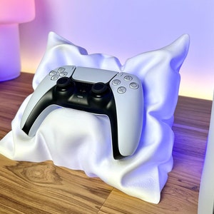 May include: A white and black PlayStation 5 controller rests on a white, cloud-shaped stand. The stand is on a wooden surface, with a white console visible in the background. The controller has a sleek, modern design.