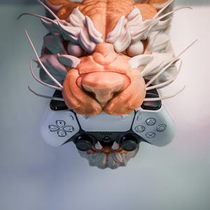 K&ouml;nnte beinhalten: Eine detaillierte Skulptur eines Tigerkopfes in Orange-, Wei&szlig;- und Graut&ouml;nen ist &uuml;ber einem wei&szlig;en und schwarzen Gamecontroller angebracht. Die Skulptur weist komplizierte Details auf, darunter Schnurrhaare und Fellstrukturen.