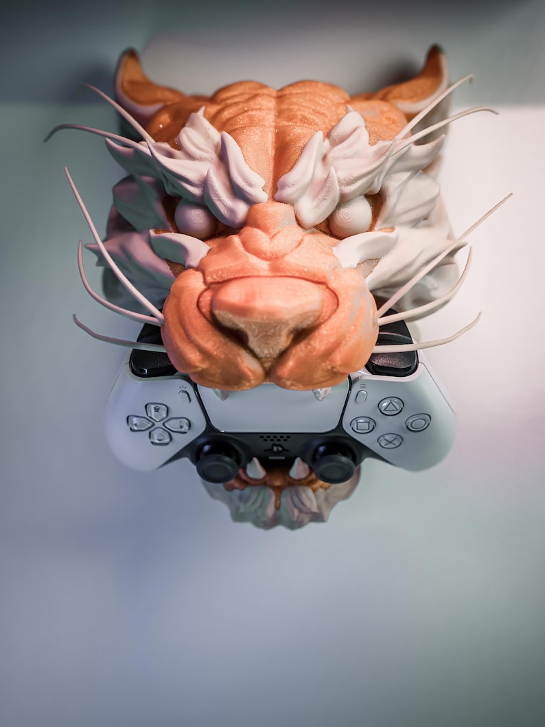 K&ouml;nnte beinhalten: Eine detaillierte Skulptur eines Tigerkopfes in Orange-, Wei&szlig;- und Graut&ouml;nen ist &uuml;ber einem wei&szlig;en und schwarzen Gamecontroller angebracht. Die Skulptur weist komplizierte Details auf, darunter Schnurrhaare und Fellstrukturen.