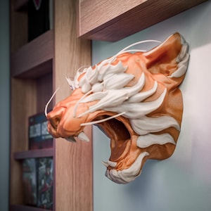 K&ouml;nnte beinhalten: Wandmontierte Tigerkopfskulptur. Die Skulptur ist in Orange und Wei&szlig; gehalten und zeigt ein detailliertes Design. Das Maul des Tigers ist ge&ouml;ffnet und zeigt Z&auml;hne. Die Skulptur befindet sich an einer hellblauen Wand neben einem Holzregal.