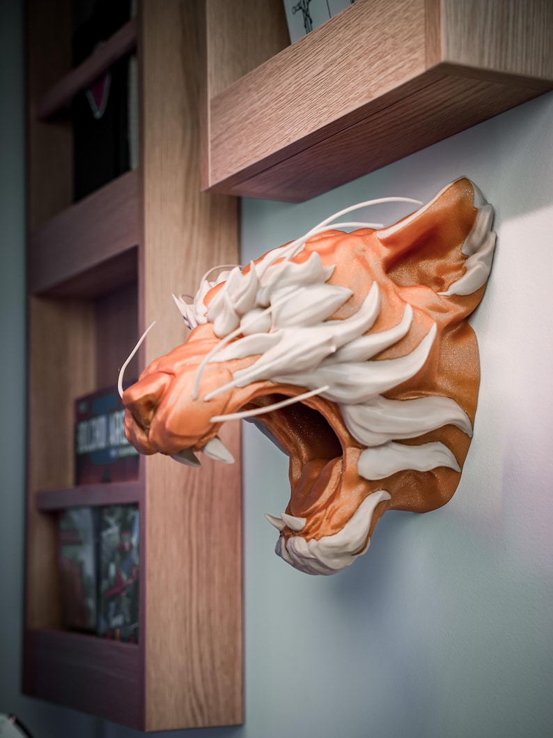 K&ouml;nnte beinhalten: Wandmontierte Tigerkopfskulptur. Die Skulptur ist in Orange und Wei&szlig; gehalten und zeigt ein detailliertes Design. Das Maul des Tigers ist ge&ouml;ffnet und zeigt Z&auml;hne. Die Skulptur befindet sich an einer hellblauen Wand neben einem Holzregal.