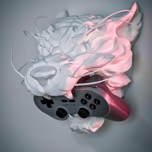 K&ouml;nnte beinhalten: Eine wei&szlig;e Skulptur eines Drachenkopfes, der einen grau-rosa Videospiel-Controller h&auml;lt. Die Skulptur hat detaillierte Texturen und wird mit rosa und wei&szlig;em Licht beleuchtet.